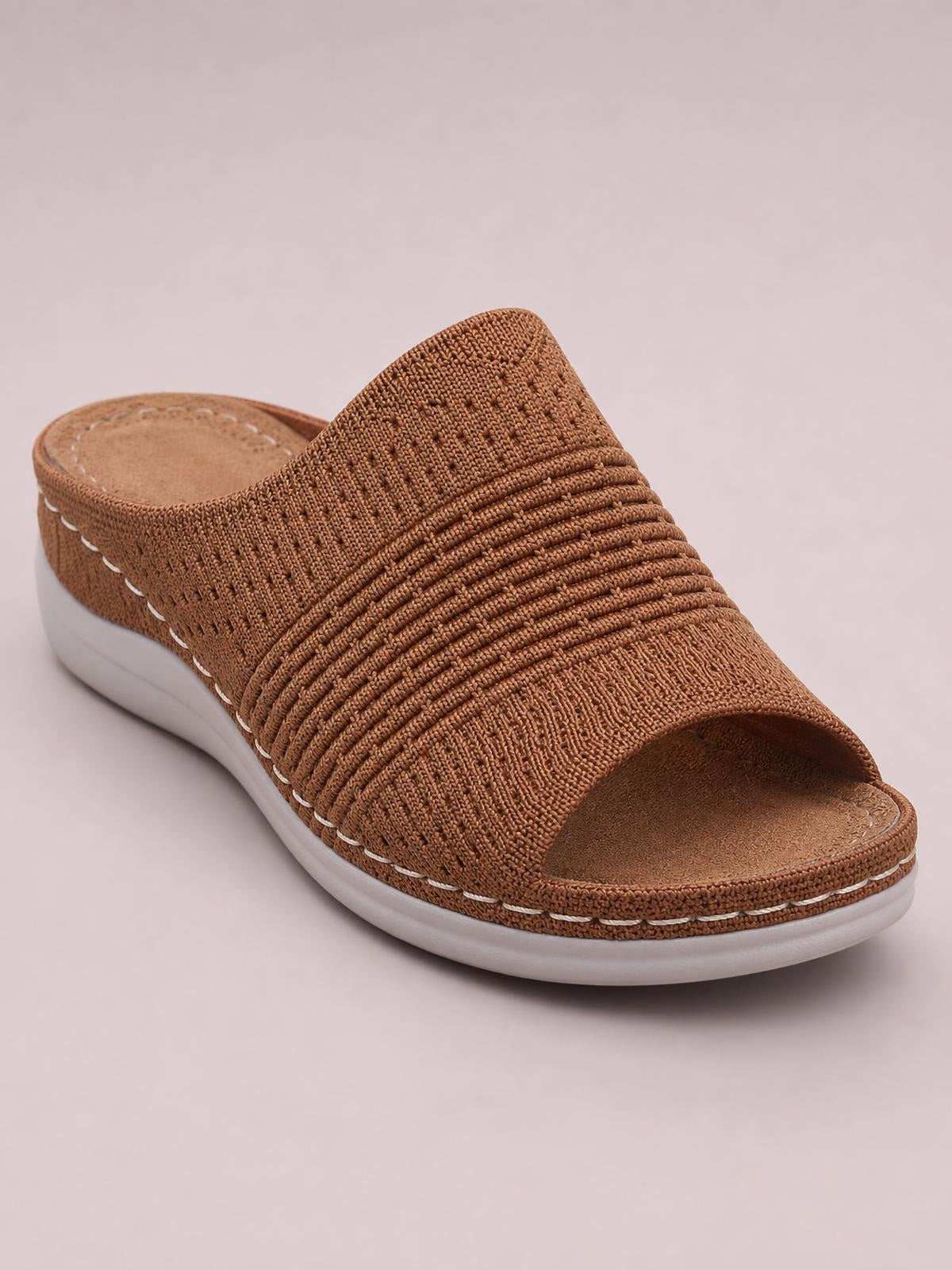 A brown mesh open-toe flat sandal features a wide textured woven upper with layered ridges, an open back, and a white rubber sole.