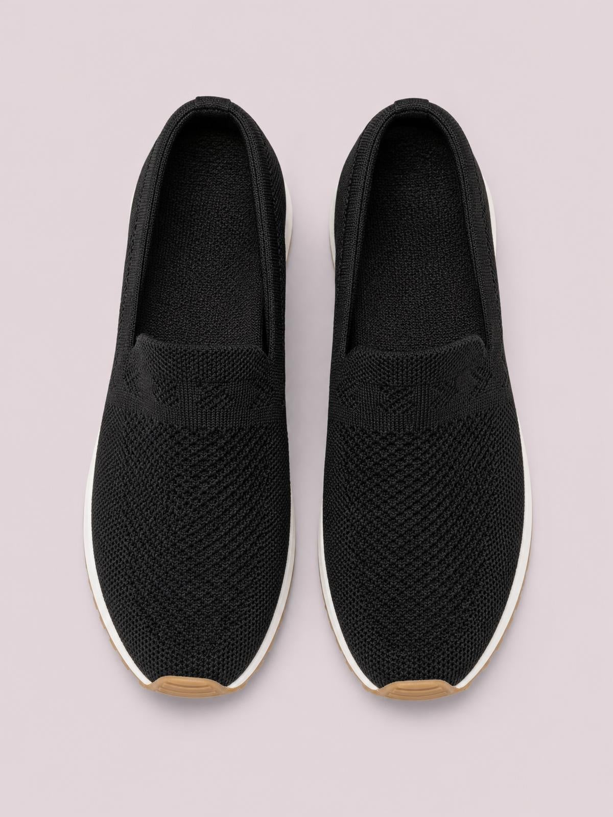 Pair of black mesh round-toe flat slip-ons feature a textured knit upper, easy slip-on opening, and contrasting white rubber soles with tan toe accents.