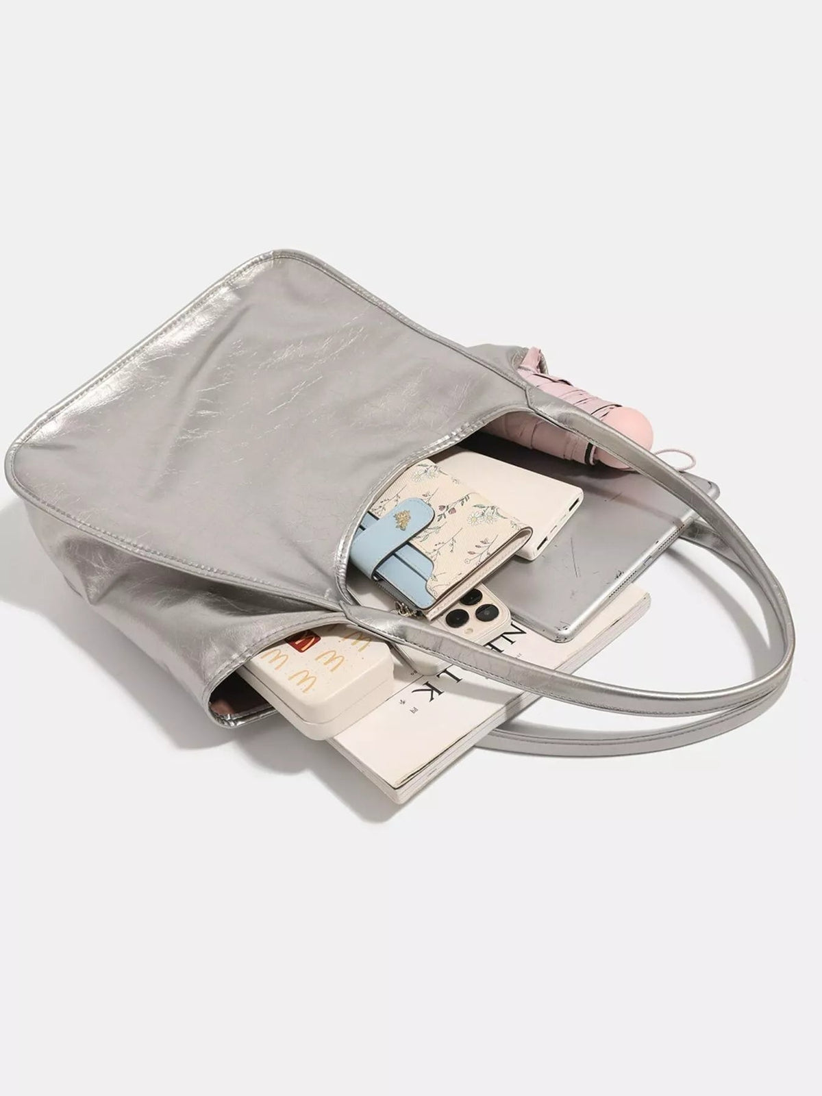 A medium metallic silver PU leather tote bag lies open, showing its spacious interior, stitched edges, long twin shoulder straps, and softly structured silhouette.