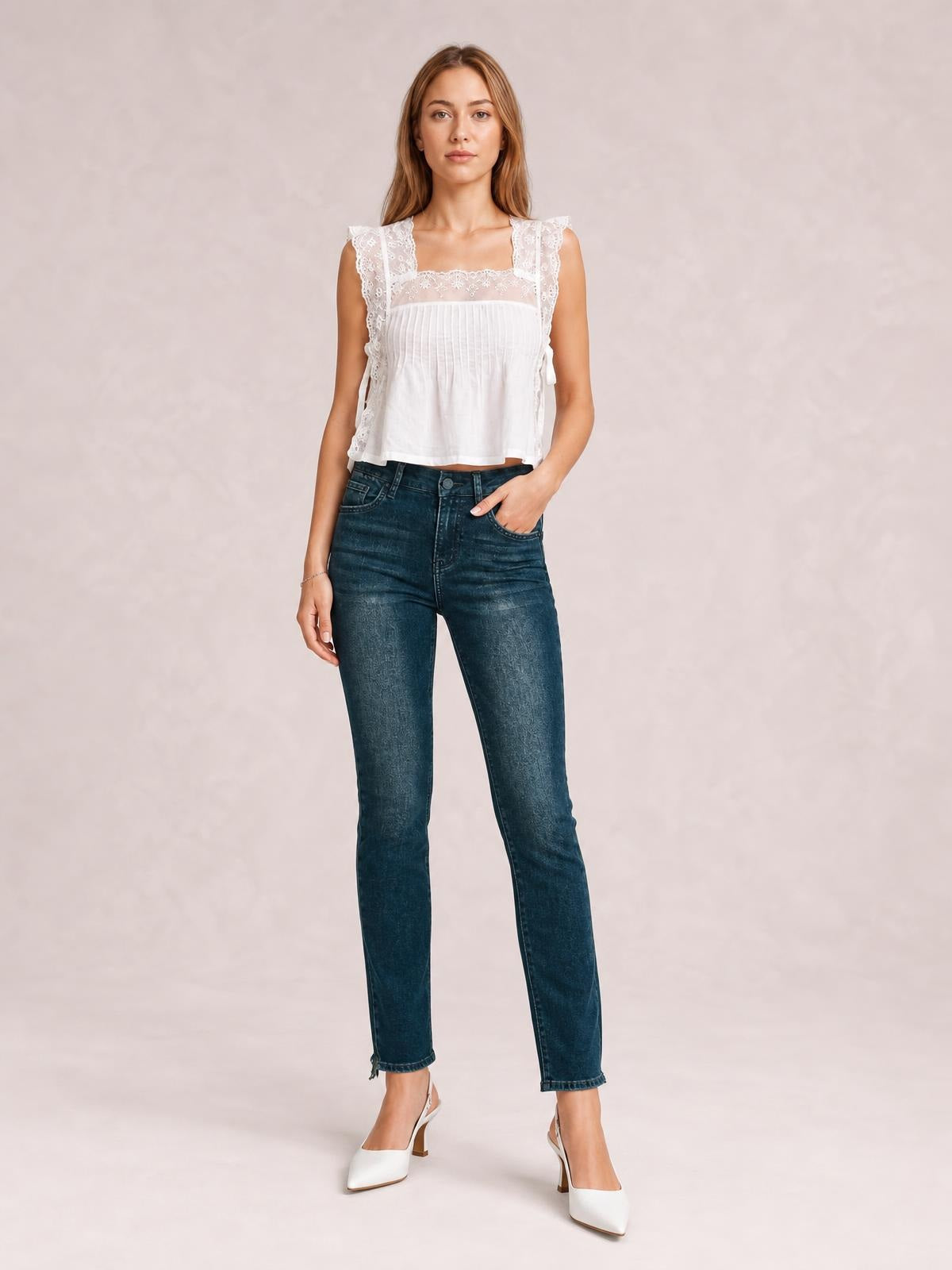 A model stands in dark blue mid-rise skinny jeans with front pockets, belt loops, button closure, subtle fading, and a slim ankle-length silhouette.