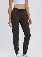 Model wears black tied joggers with a drawstring waist, side pockets, tapered legs, and fitted ankle cuffs in a relaxed standing pose.