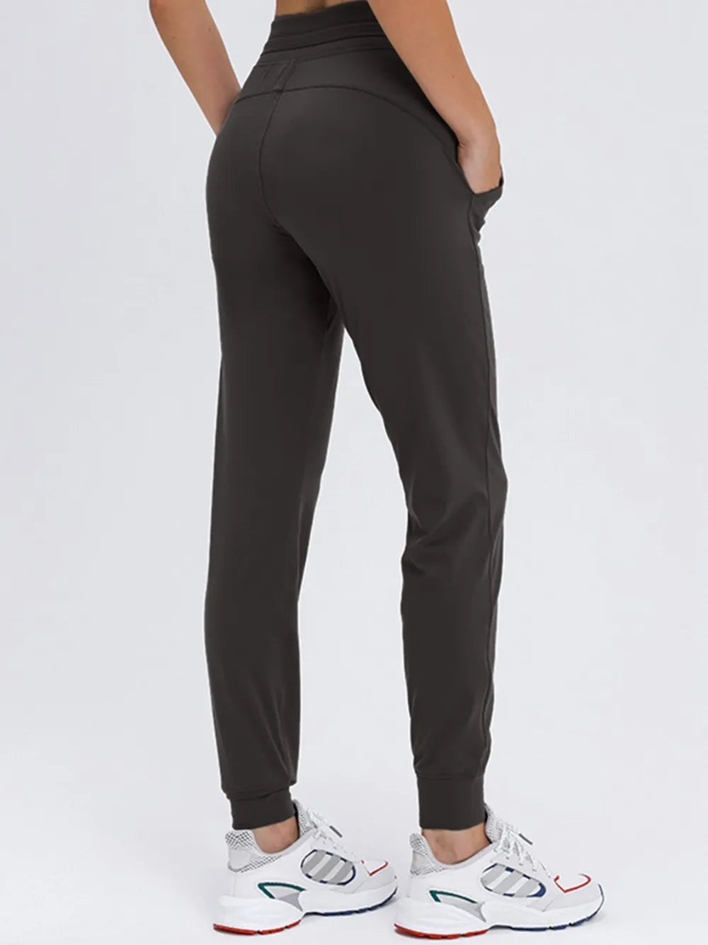 Model shown from behind wears black tied joggers with a smooth waistband, curved seam detailing, side pockets, tapered legs, and ribbed ankle cuffs.