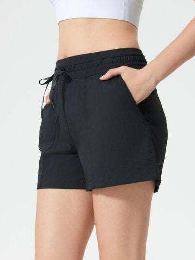 Model wearing black drawstring active shorts with an elastic waistband, side pockets, and a relaxed silhouette, posing with one hand tucked into a pocket.