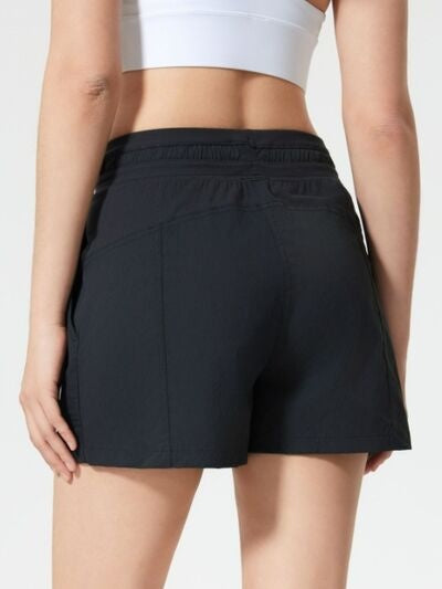 A model stands with hands at her sides, wearing black drawstring active shorts with an elastic waistband, side pockets, and a relaxed mid-thigh silhouette.