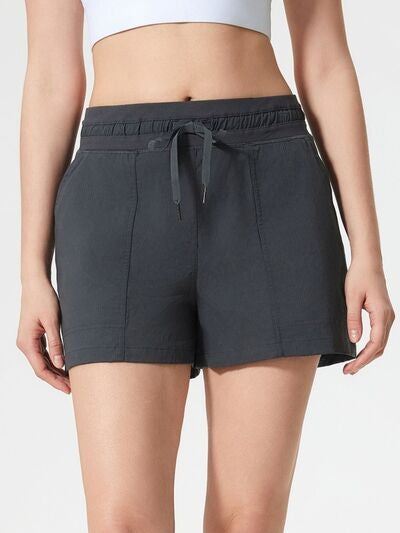Model wears dark charcoal active shorts with an elastic drawstring waistband, visible front pockets, and a relaxed straight-leg silhouette ending above the knee.