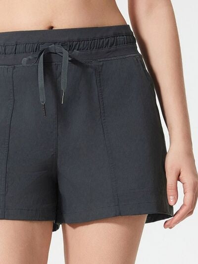 A model wears dark gray drawstring active shorts with an elastic waistband, front pockets, moderate stretch, and a relaxed straight-leg silhouette.