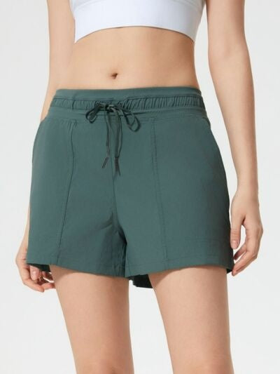 A standing model wears dark teal drawstring active shorts with an elastic waistband, relaxed straight-leg silhouette, and visible side pockets.