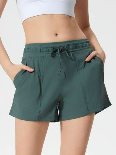 A model wears dark teal drawstring active shorts with an elastic waist, relaxed straight-leg silhouette, visible stitching, and hands tucked into the pockets.