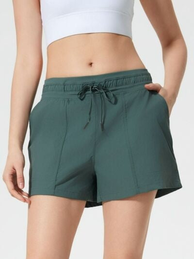 Model wearing dark teal drawstring active shorts with side pockets, an elasticized waistband, relaxed straight-leg silhouette, and moderate stretch.