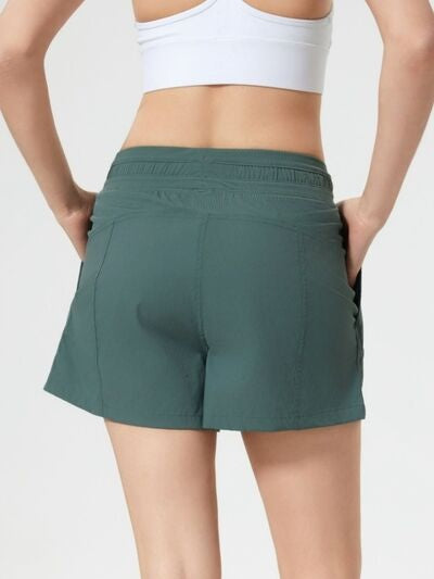 A model shown from behind wears dark green drawstring active shorts with a relaxed mid-thigh silhouette, elastic waistband, and visible side pockets while holding the waistband.