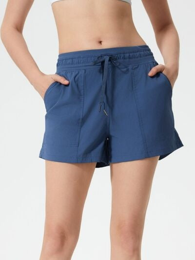 Model wears blue drawstring active shorts with an elastic waistband, relaxed silhouette, side pockets, and a lightweight, moderately stretchy construction.