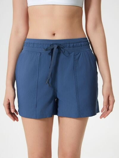 Visible model wears navy blue drawstring active shorts with an elastic waistband, side pockets, moderate stretch, and a relaxed straight-leg silhouette.