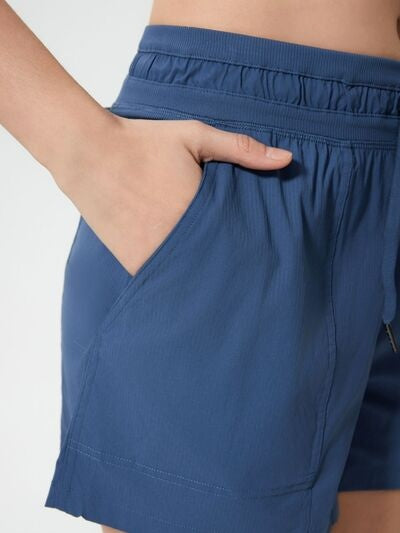 A model wears navy blue drawstring active shorts with an elastic waistband, angled side pocket, relaxed straight-leg silhouette, and stitched hem.