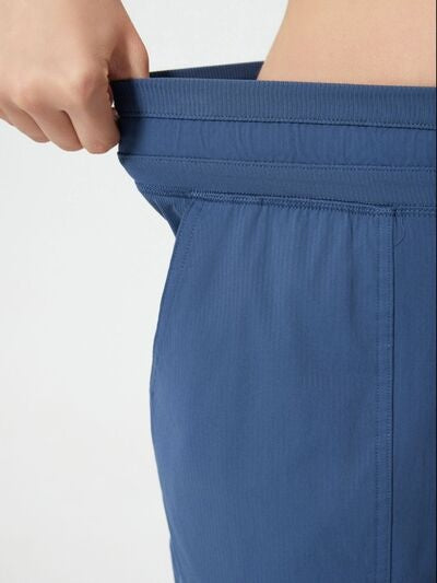 Close-up of blue drawstring active shorts shows a hand pulling the elastic waistband, with stitched construction and a relaxed, comfortable silhouette.