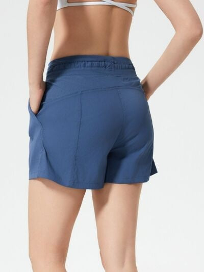Back view of a model wearing blue drawstring active shorts with side pockets, an elastic waistband, moderate stretch, and a relaxed athletic silhouette.