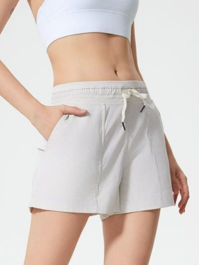 Model wears light gray drawstring active shorts with an elastic waistband, side pockets, and a relaxed straight-leg silhouette, shown with one hand tucked into a pocket.