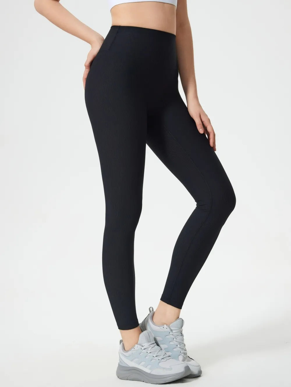 Model wears black high waist active leggings with a sleek, close-fitting ankle-length silhouette, standing with one knee bent and hands resting on her hips and thigh.