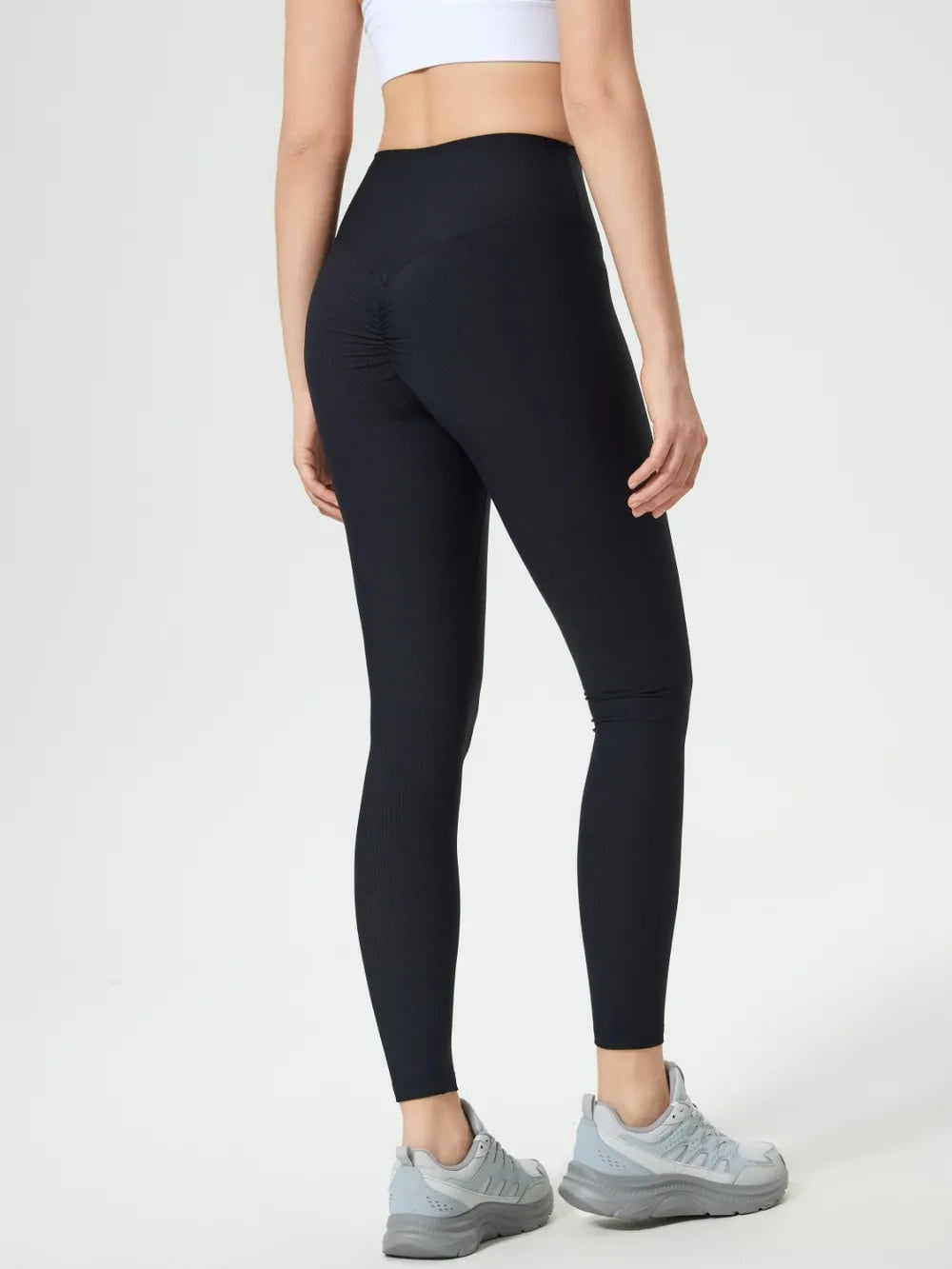 Model from behind wearing black high-waist active leggings with a fitted full-length silhouette and gathered seam detail, standing with one leg slightly bent.