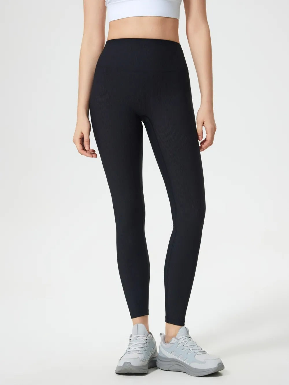 Model wears black high-waist active leggings with a wide waistband, subtle ribbed texture, stretchy slim silhouette, and ankle-length hems.