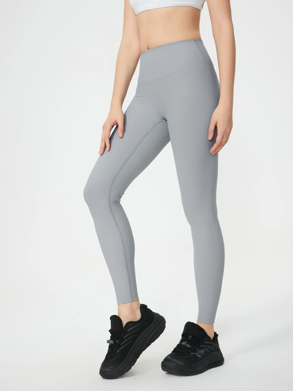 Model wearing light gray high-waist active leggings with a smooth, stretchy fitted silhouette, ankle-length legs, and one knee bent in a casual athletic pose.