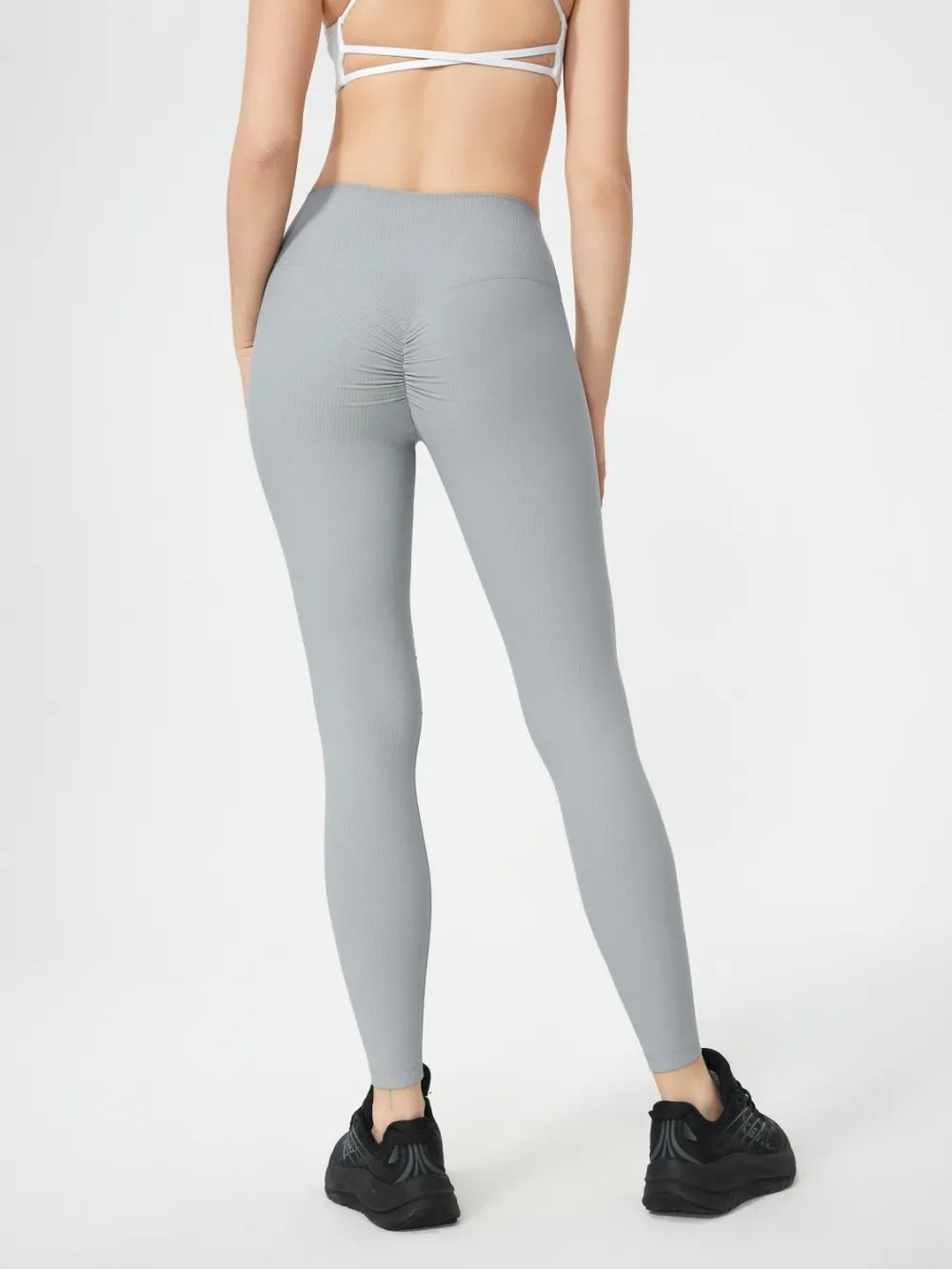 A model poses from behind in light gray high-waist active leggings with a stretchy, close-fitting silhouette, gathered seat seam, and ankle-length hems.