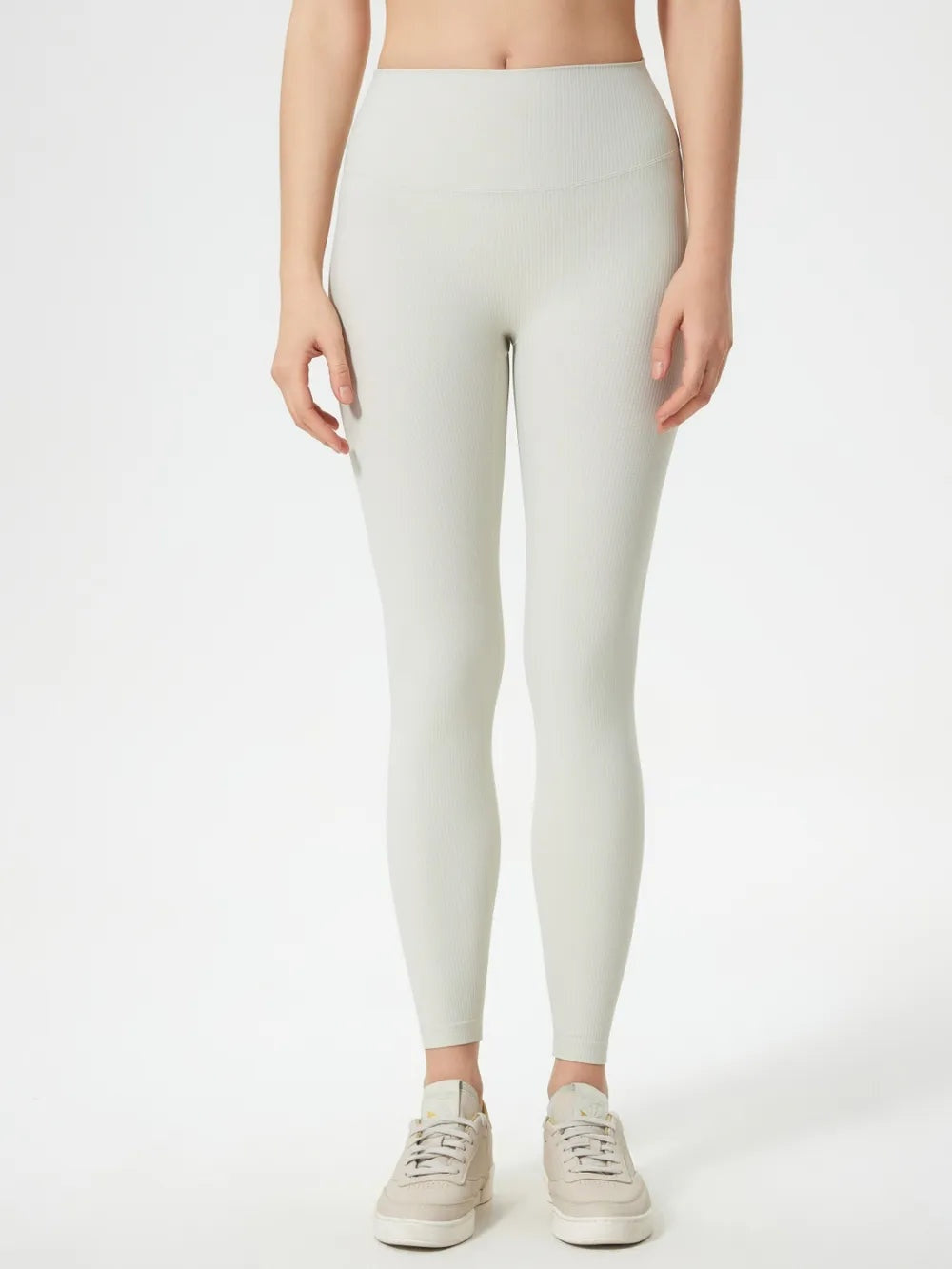 Model stands in light ivory high-waist active leggings with a close-fitting full-length silhouette, wide waistband, subtle vertical texture, and curved front seam.
