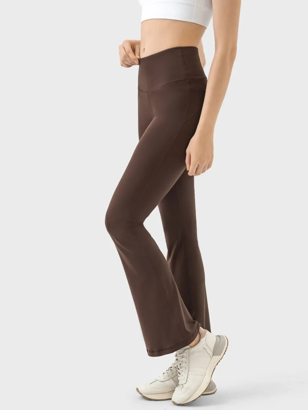 A model wears brown high-waist active pants with a wide supportive waistband, seamed construction, stretchy fitted legs, and gently flared hems while standing with one knee bent.