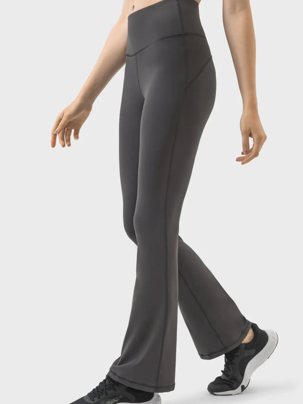 Model wearing dark gray high waist active pants with a fitted hip and thigh, flared legs, curved seam detailing, and a crossed-leg walking pose.