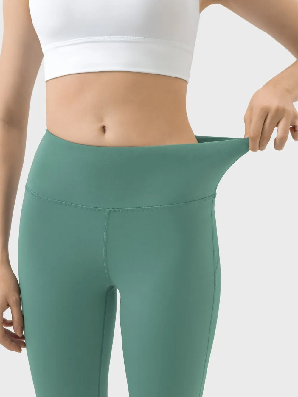 Model wearing high waist teal active pants with a wide supportive waistband, fitted stretchy silhouette, and visible seam construction while pulling the waistband outward.