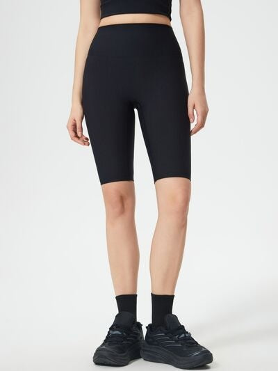 Model stands in black high waist active shorts with a fitted, above-knee bike-short silhouette and smooth stretch construction.