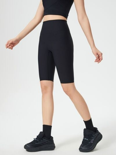 Model wears black high waist active shorts with a sleek, fitted knee-length silhouette and stretchy construction, standing with one leg crossed forward.