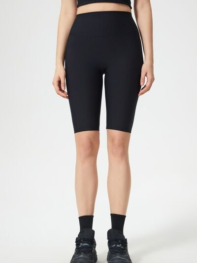 A model stands in black high waist active shorts with a smooth, fitted silhouette, stretchy construction, and a longer length ending just above the knees.