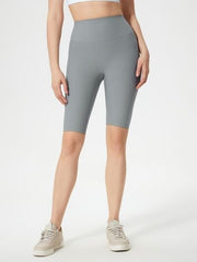 Model wears gray high waist active shorts with a smooth, fitted knee-length silhouette and stretchy construction, standing with one leg slightly forward.