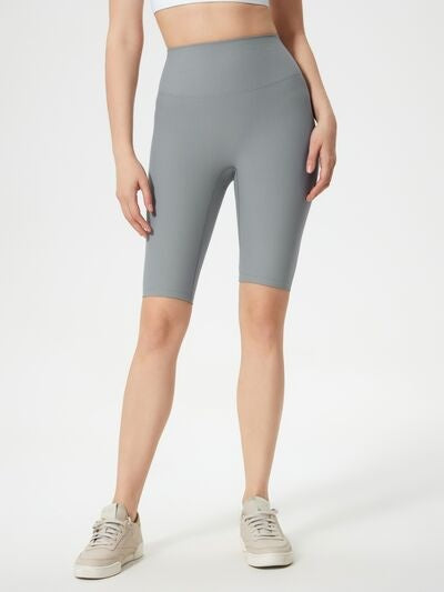 Model wears gray high waist active shorts with a smooth, fitted knee-length silhouette and stretchy construction, standing with one leg slightly forward.