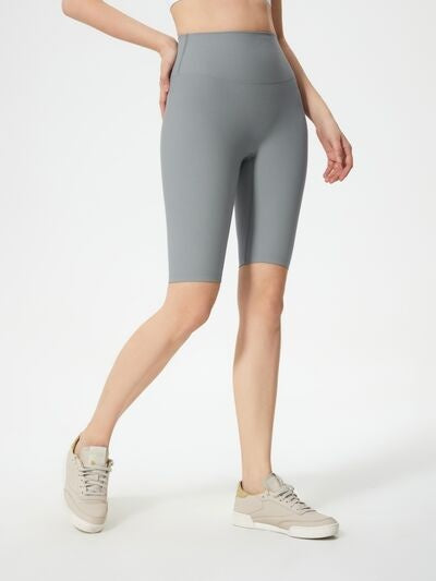 Model wears gray high-waist active shorts with a smooth, fitted knee-length silhouette, standing with one leg crossed forward for a sleek athletic look.