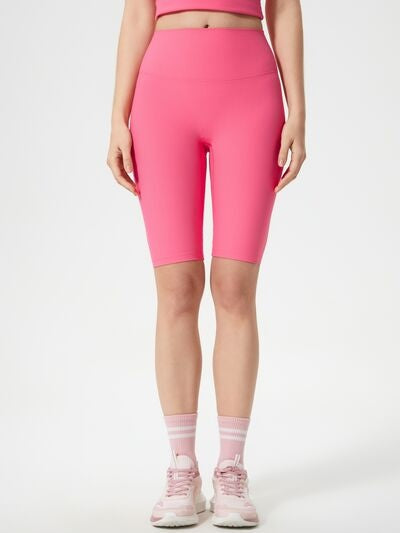 A model stands wearing bright pink high waist active shorts with a wide waistband, fitted silhouette, and knee-length stretch construction.