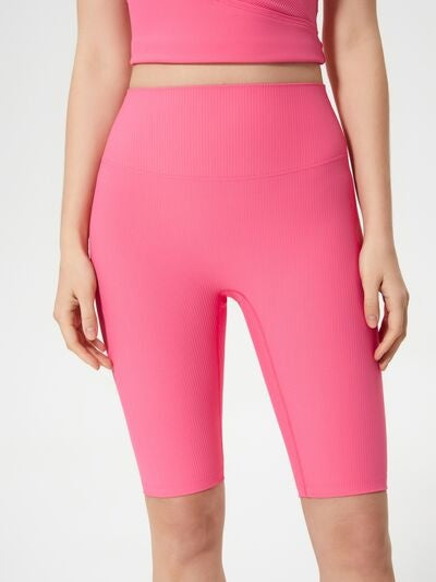 Model wearing bright pink high waist active shorts with a fitted knee-length silhouette, stretchy ribbed construction, and hands resting at her sides.