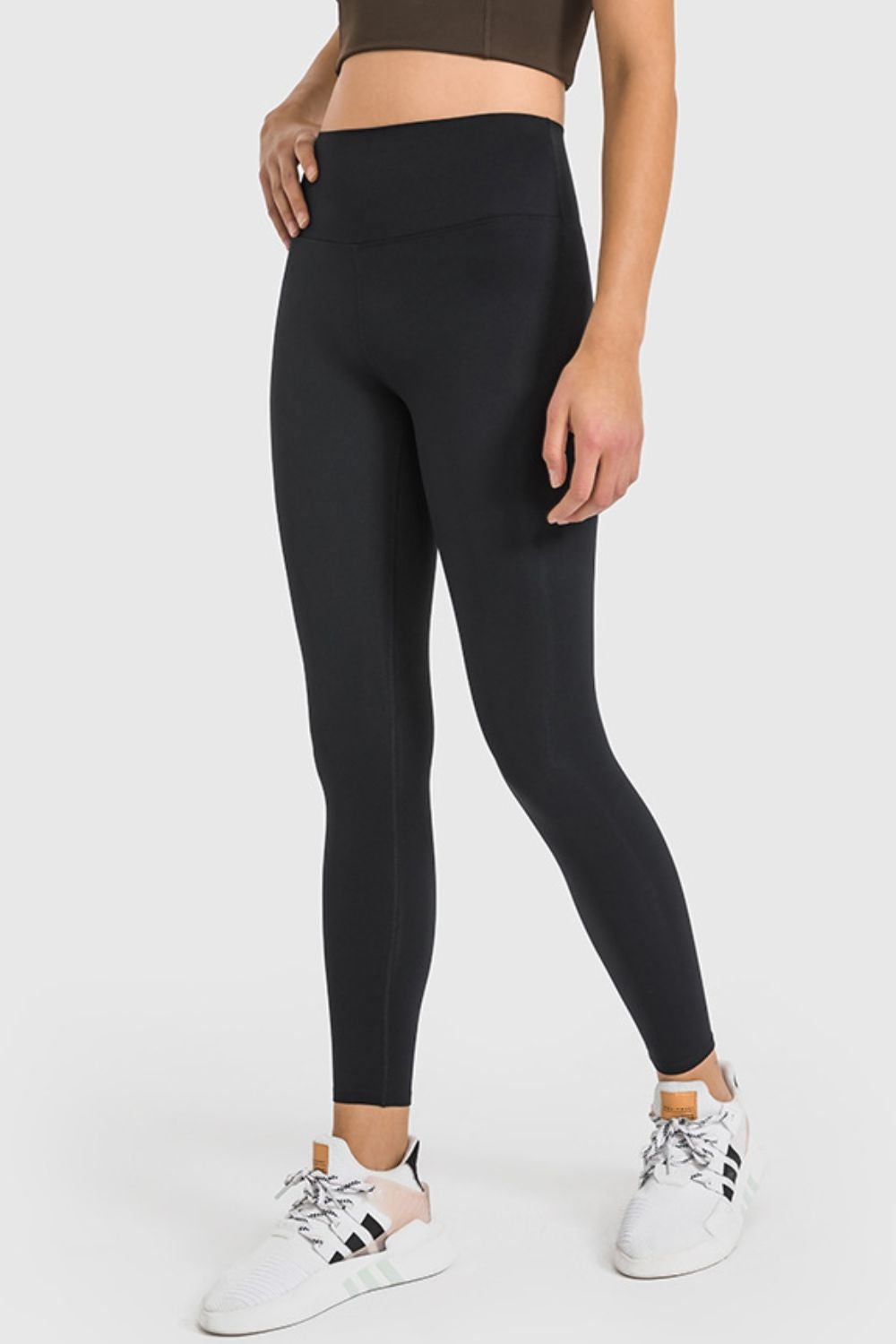 Model poses in black high-waist ankle-length yoga leggings with a wide waistband, sleek fitted silhouette, and visible leg seams.