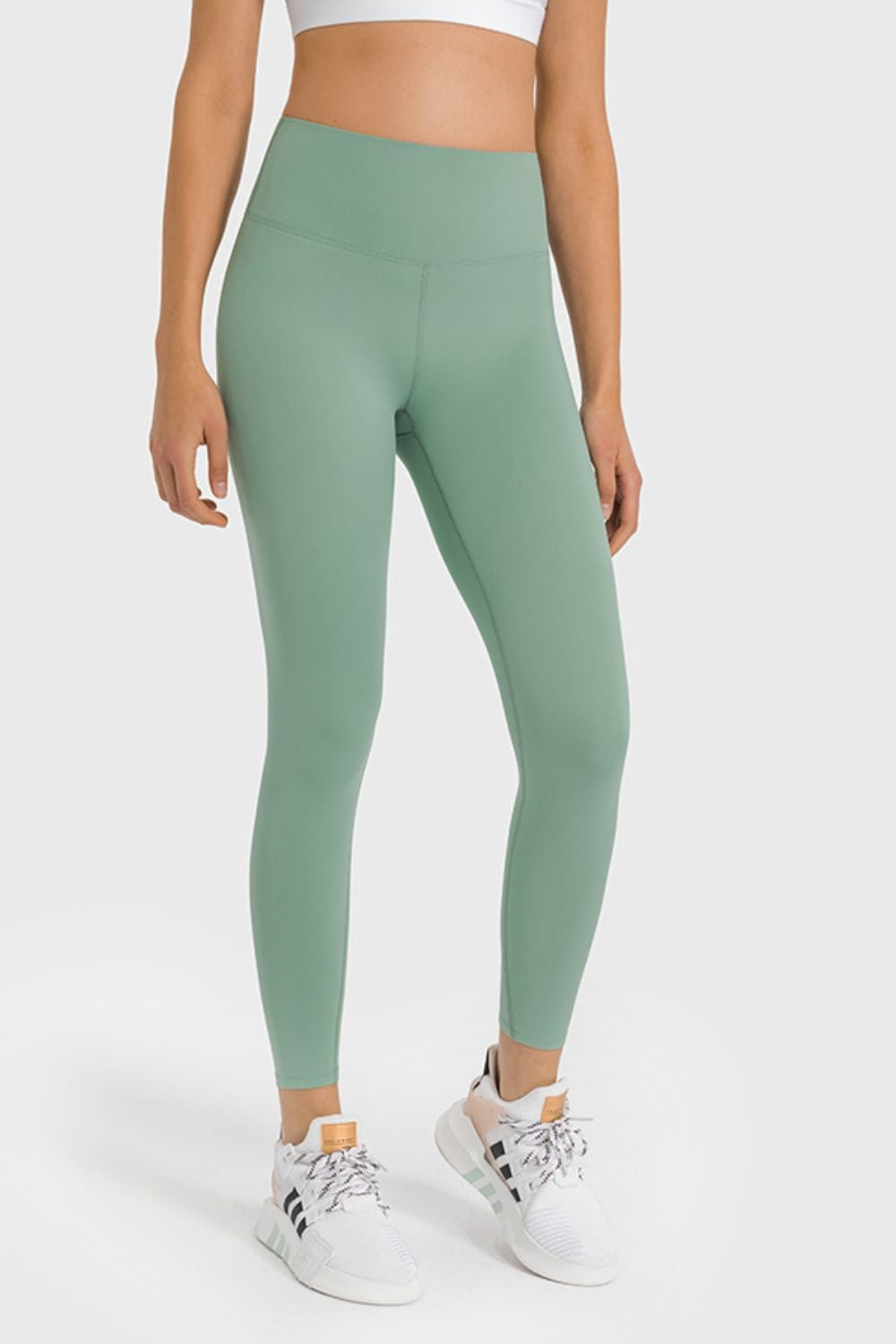 Model wears sage green high-waist ankle-length yoga leggings with a smooth, stretchy, fitted silhouette and visible front crotch seam.