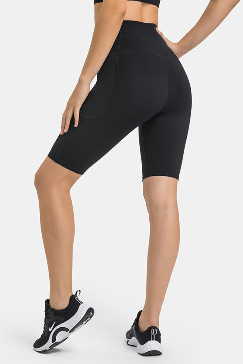 Model poses in black high-waist biker shorts with a fitted, stretchy knee-length silhouette, curved panel seams, and visible side pockets.
