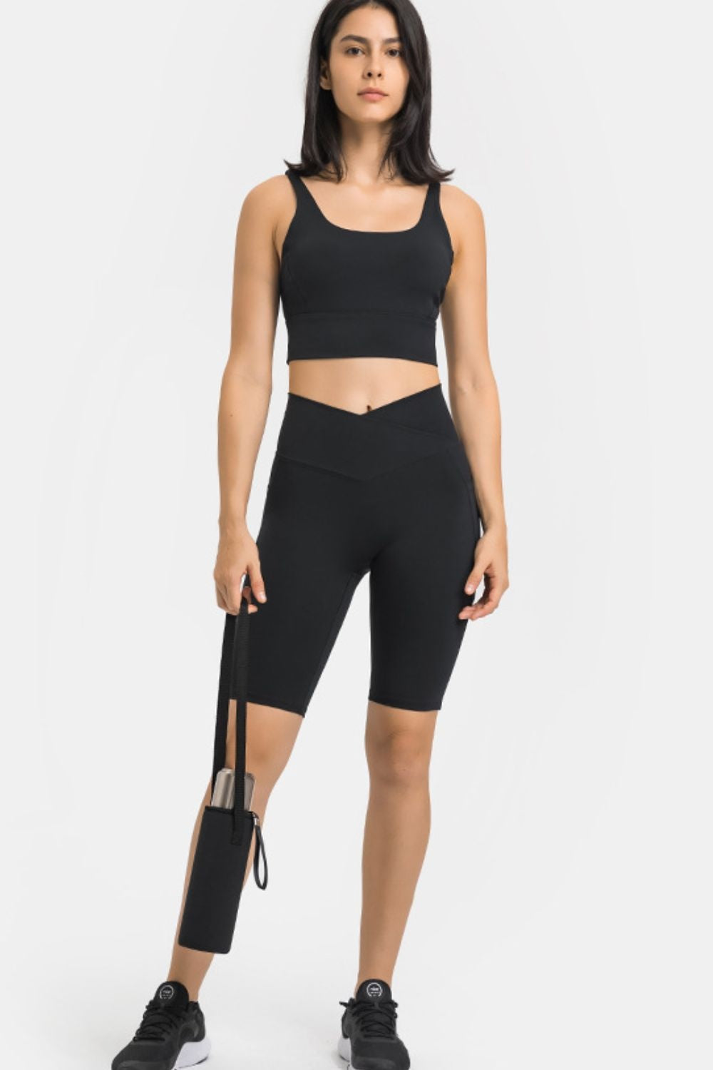 A woman stands facing forward in fitted black high-waist biker shorts with a crossover waistband and side thigh pocket, paired with a black crop top.