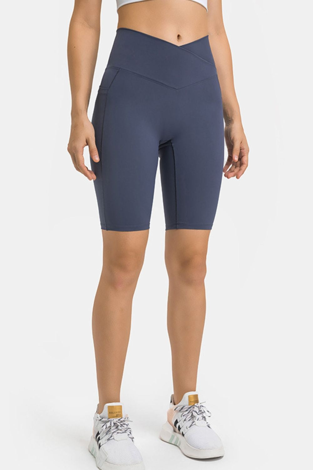Standing model wears navy high-waist biker shorts with a V-shaped waistband, fitted knee-length silhouette, visible side pocket, and smooth seamed construction.