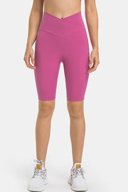 Model stands facing forward in bright pink high-waist biker shorts with a crossover waistband, fitted stretch silhouette, and knee-length construction.