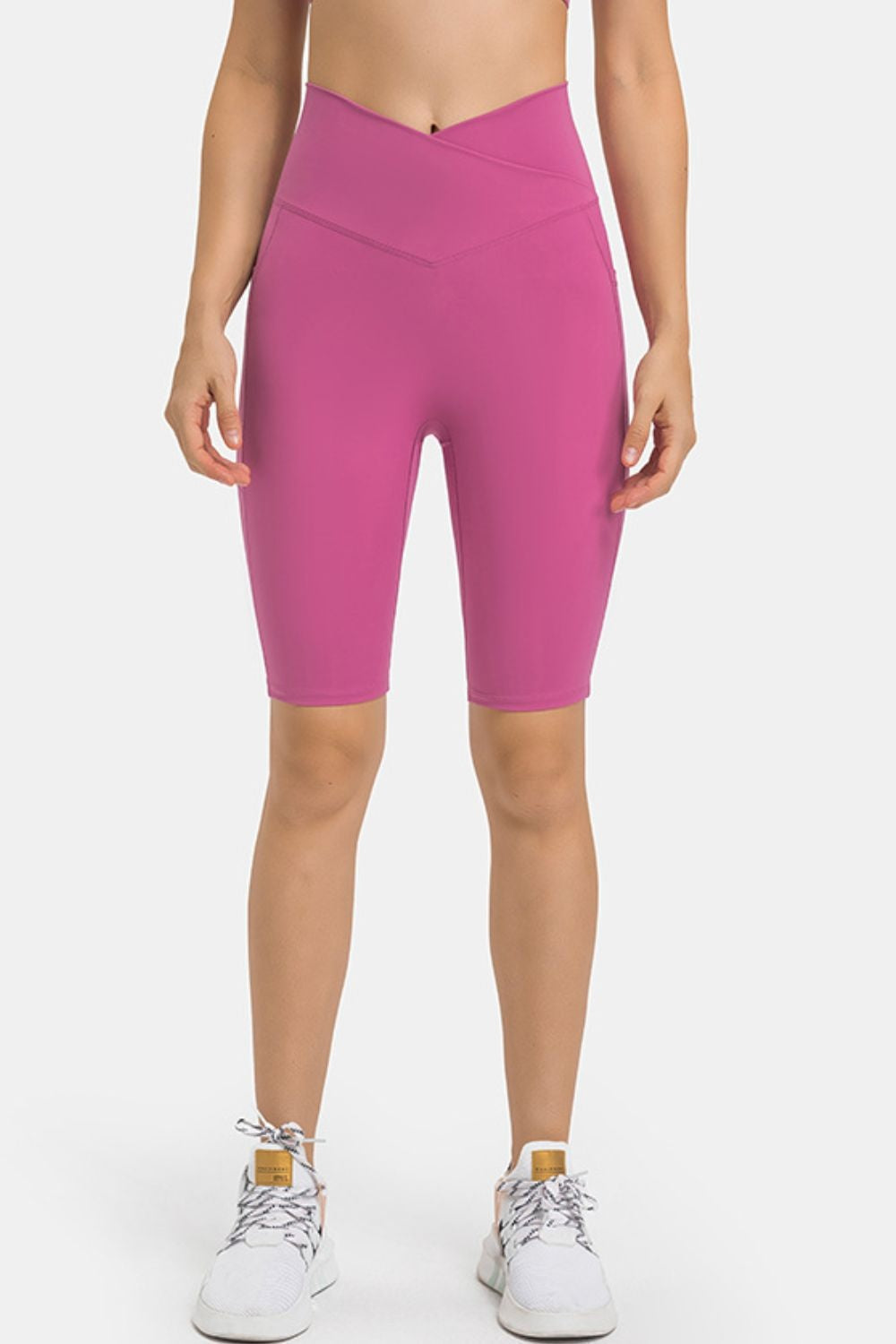 Model stands facing forward in bright pink high-waist biker shorts with a crossover waistband, fitted stretch silhouette, and knee-length construction.
