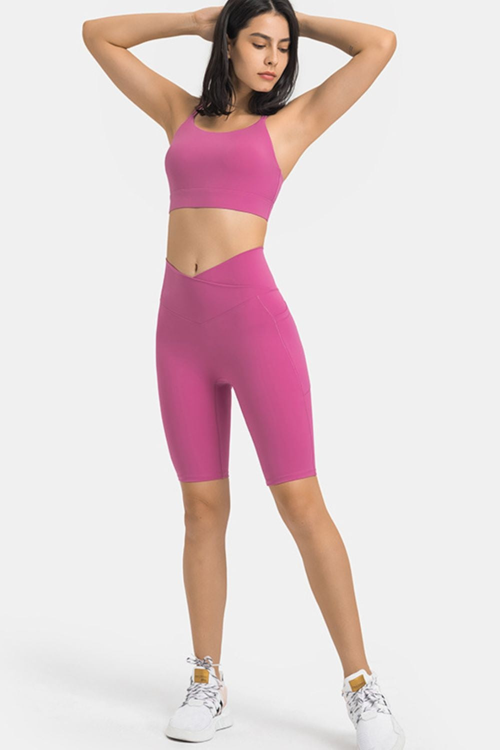 A model poses with her arms behind her head, wearing pink high-waist biker shorts with side pockets, a fitted knee-length silhouette, and stretch construction.