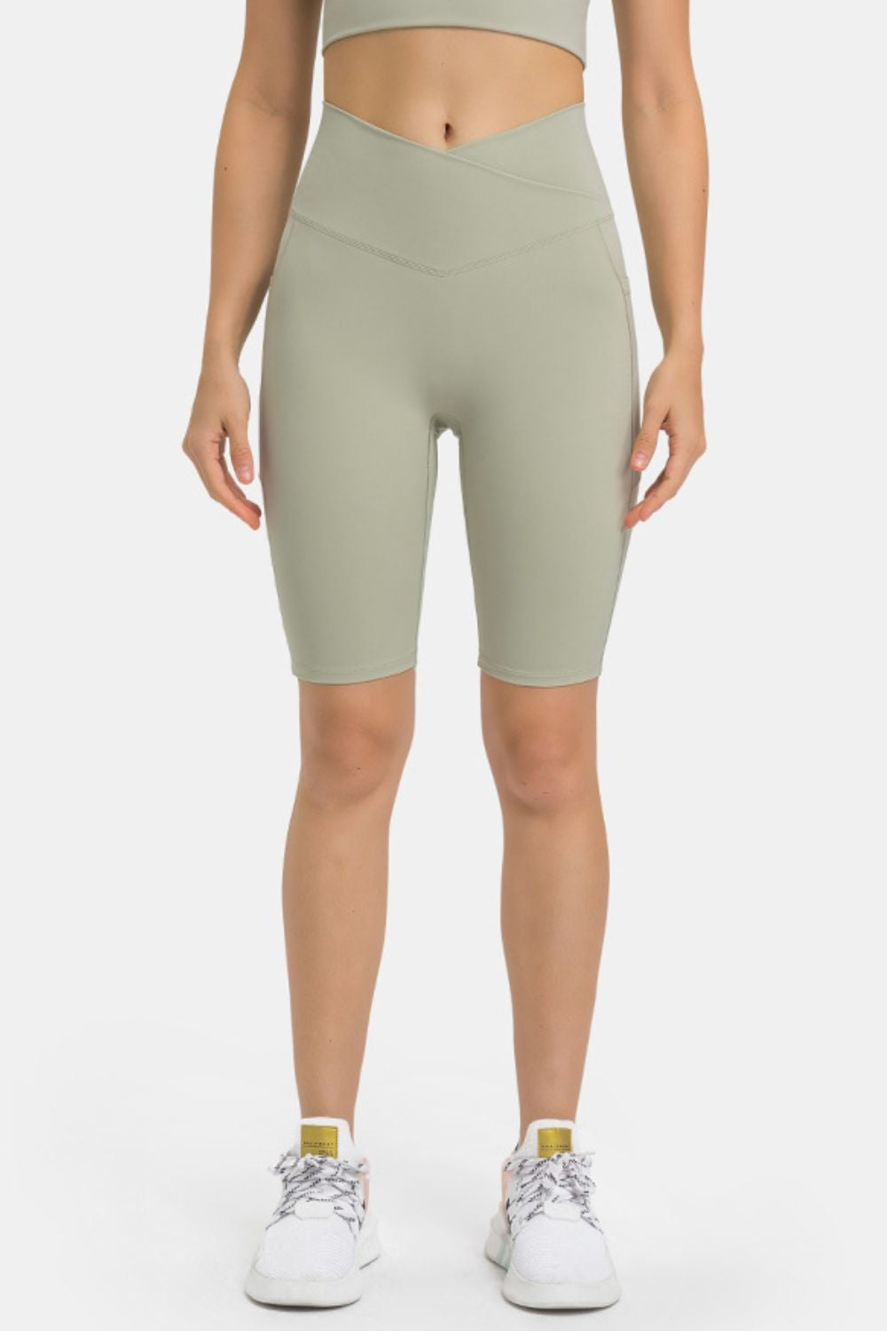 Model standing in sage green high-waist biker shorts with a fitted knee-length silhouette, stretchy construction, and overlapping V-shaped waistband seams.