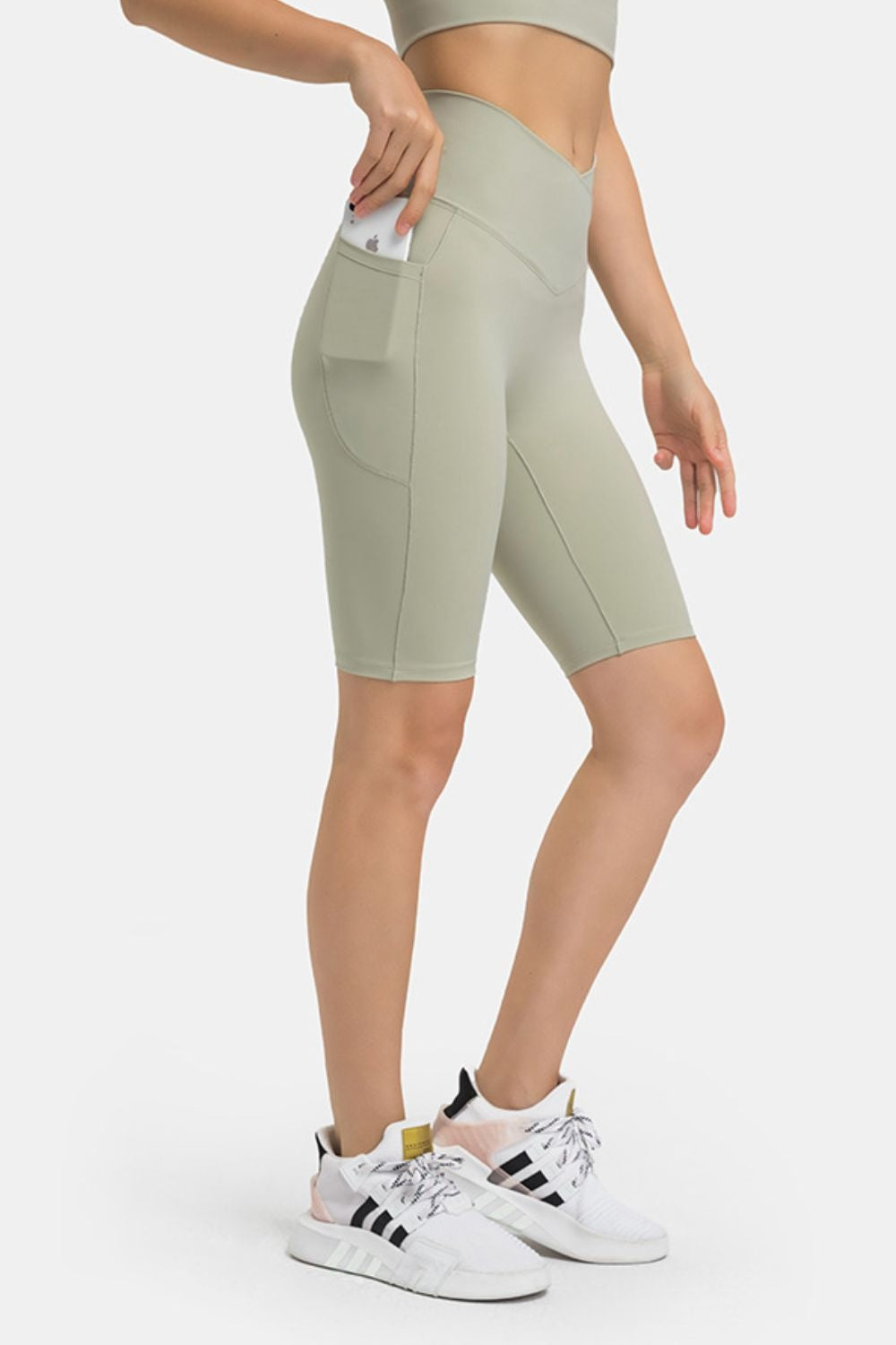 Model wears sage green high-waist biker shorts with side pockets, stitched panel construction, and a fitted knee-length silhouette while posing with one hand in a pocket.