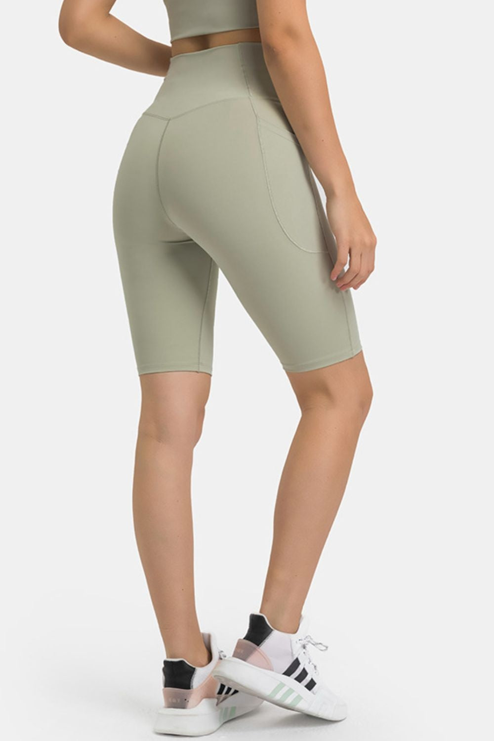 Model wearing sage green high-waist biker shorts with side pockets, a fitted knee-length silhouette, curved seam construction, and one hand on her hip.