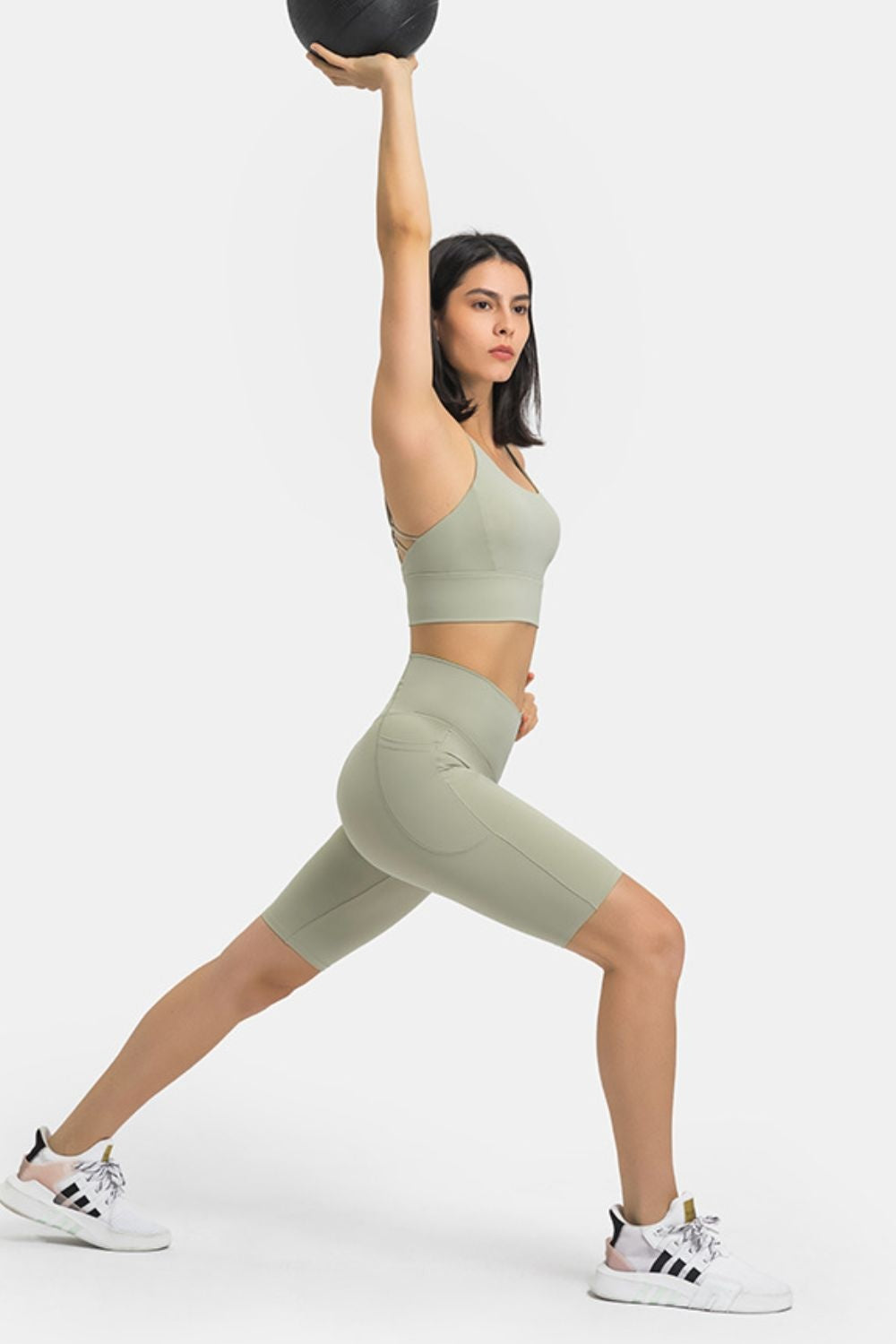 A model lunges in sage green high-waisted biker shorts with a visible side pocket, fitted stretchy silhouette, and clean seamed construction.