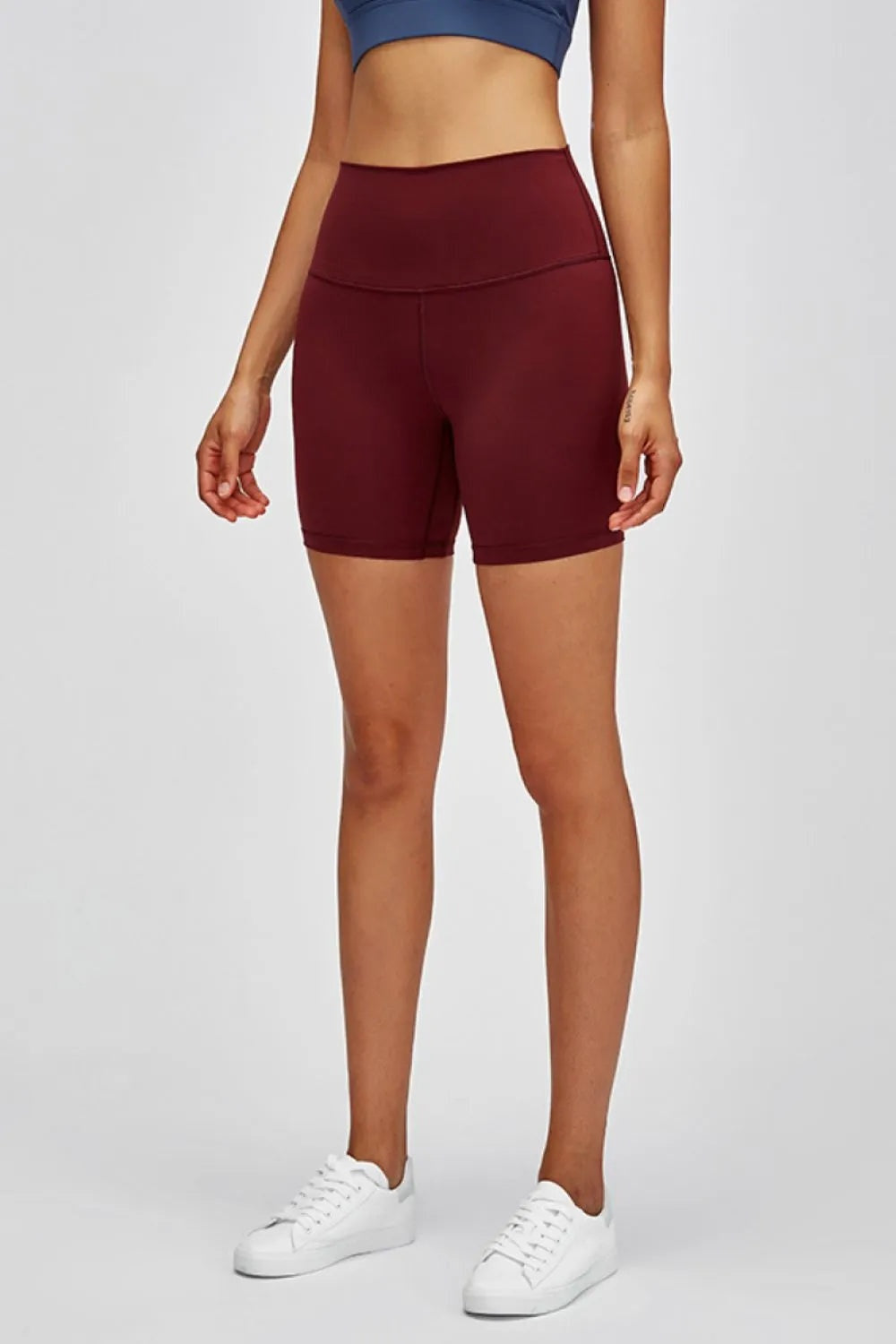 Model standing in burgundy high-waist training shorts with a fitted stretch silhouette, wide waistband, curved seam, and above-knee length.